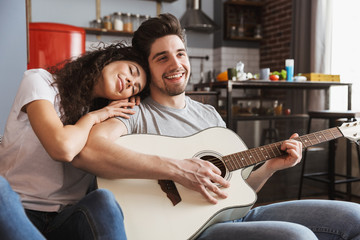 Image of happy young couple sitting on sofa at home and playing music on acoustic guitar