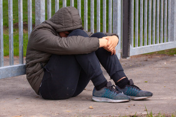 Lonely teenager sitting alone outside a metal fence with iron bars. Loneliness and bullying concept.