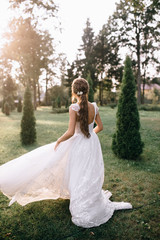 Fototapeta na wymiar back view of beautiful young bride in white wedding dress standing on green lawn at sunset, backlit