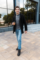 Portrait of masculine man holding paper cup of takeaway coffee while strolling in front of glass building