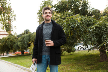 Photo of unshaved man holding paper cup of takeaway coffee while walking through green park