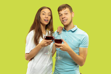 Beautiful young couple's half-length portrait isolated on green studio background. Woman and man standing with glasses of red wine. Facial expression, summer, weekend concept. Trendy colors.