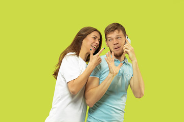 Beautiful young couple's half-length portrait isolated on green studio background. Man speaking on the phone, woman is angry for it. Facial expression, human emotions concept. Trendy colors.