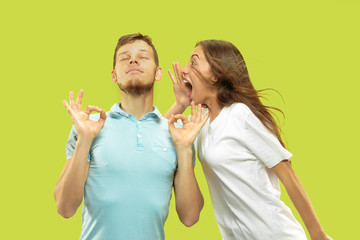 Beautiful young couple's half-length portrait isolated on green studio background. Man is trying to keep calm with closed eyes while woman is screaming. Facial expression, human emotions concept.