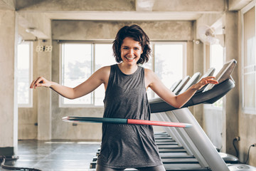 Beautiful young smiling happy Caucasian sporty woman with short hair playing hula hoop inside gym studio with treadmills behind - fun workout fitness portrait concept - obrazy, fototapety, plakaty