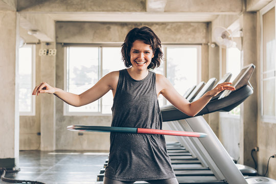 Beautiful young smiling happy Caucasian sporty woman with short hair playing hula hoop inside gym studio with treadmills behind - fun workout fitness portrait concept