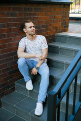 A young handsome man with bristles sits on a granite staircase near a brick building on a summer evening