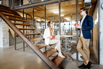 Fototapeta na wymiar Young buiness man and woman having a conversation, sitting on the stairs during the coffee break in the modern office, wide interior view