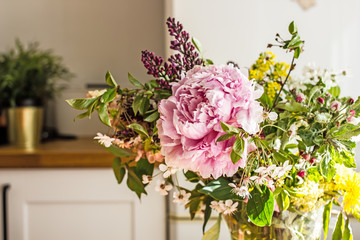 A spring bouquet of pink peony flowers, a blossoming apple tree branch, a lilac flower and wild flowers in a transparent jug are in the kitchen. Copy space.