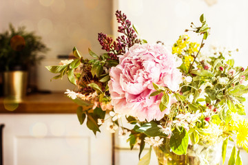 A spring bouquet of pink peony flowers, a blossoming apple tree branch, a lilac flower and wild flowers in a transparent jug are in the kitchen. Copy space.
