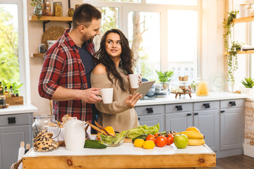 Beautiful young couple is talking,  smiling while eating tea or coffee and drinking in kitchen at home. Using tablet.
