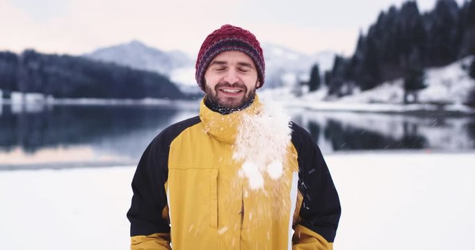 Portrait of charismatic man in a orange jacket in front of the camera smiling large someone shoot the snow ball in his hat funny moments outside in the middle of landscape with lake and snowy mountain