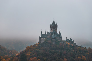 Reichsburg Cochem / Castle Cochem covered in fog during an orange and misty autumn day with low hanging clouds and fog (Cochem, Germany, Europe)