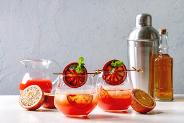 Blood orange iced cocktails in glasses, decorated by slice of oranges and fresh mint on skewers, served with shaker, jug of juice, bottle of rum on white marble table with grey wall at background.