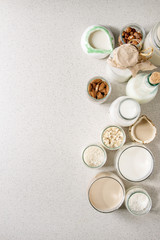 Obraz na płótnie Canvas Variety of non-dairy vegan lactose free nuts and grain milk almond, hazelnut, coconut, rice, oat in glass bottles and ceramic jugs with ingredients above over white spotted background. Flat lay, space