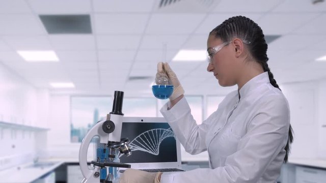 A female lab technician sitting at a table next to a laptop in a modern medical laboratory examines a sample of blue liquid in a glass flask
