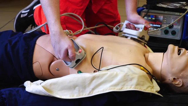 Close up static shot on unrecognisable male paramedic in orange scrubs teaching students correct procedure on using defibrillator machine on dummy patient.