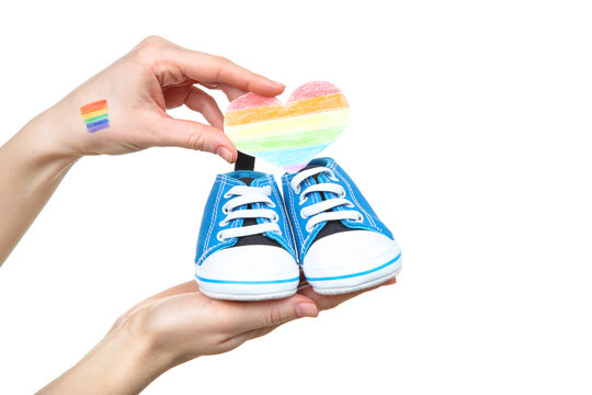 Female hand with drawing rainbow flag, paper heart and baby shoes on white background