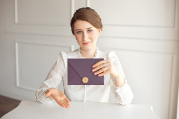 girl in a white business style shirt with a classic hairdo on brunette hair holding a welcome envelope in her hand sitting at a table in a white background.