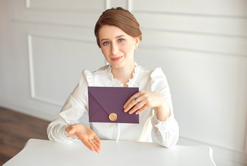 girl in a white business style shirt with a classic hairdo on brunette hair holding a welcome envelope in her hand sitting at a table in a white background.