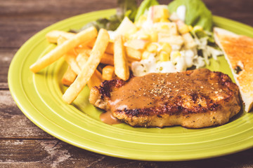 Pig steak is a fast food,pig steak this dish consists of with salad which has  purple lettuce corn apple cabbage salad dressing bread french fries and black pepper sauce, all in a green plate.