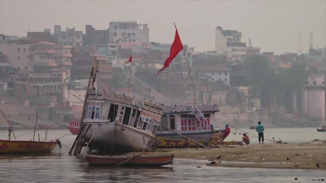 Medium low-angle still shot facing Ganga riverbank of crudely anchored, and abandoned local boats at a dirt ground. Close by are Indian men, Varanasi, Ganga River, India