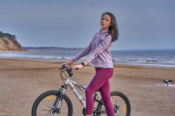 Fototapeta na wymiar Young slim brunette woman on a bike ride on the sandy shore of a large river. Cloudy summer day.