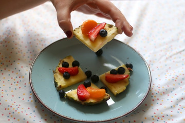 Various spring fruit on a turquoise plate: strawberries, pineapple, blueberries and tangerine. Unrecognizable person reaching for fruit. Selective focus.