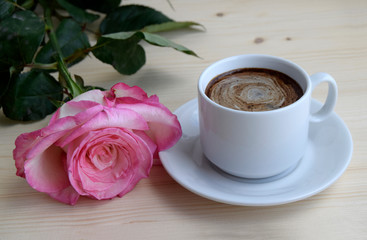 The cup of coffee. Pink rose flower. The flower and cup of coffee are on a wooden surface.