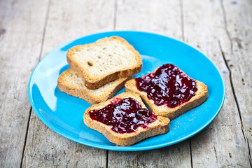 Fresh toasted cereal bread slices with homemade wild berries jam on blue ceramic plate closeup on rustic wooden background.