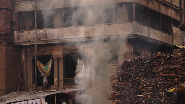 Close-up low-angle still shot of an old Varanasi city building with white cremation smoke rising close to a pile of firewood, Markarnika ghat, Ganga River, India