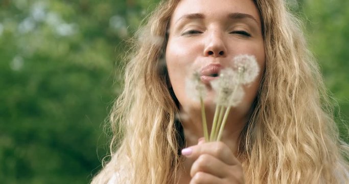 Adult woman blowing on the dandelions on the nature. Happy adult  girl blows dandelions. Happiness concept. Outdoors portrait of a blonde woman with long curly hair in the park. Smiling female face