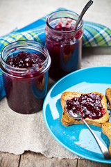 Fresh cereal bread slices on blue ceramic plate, homemade cherry and wild berries jam in jars and spoons closeup on linen napkin on rustic wooden table background.
