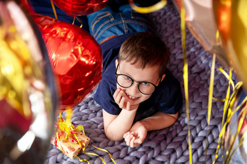 happy boy with glasses laughing lying on the bed, holiday decorated with balloons