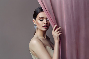 A beautiful, tender, romantic girl stands behind a transparent curtain in elegant pearl earrings...