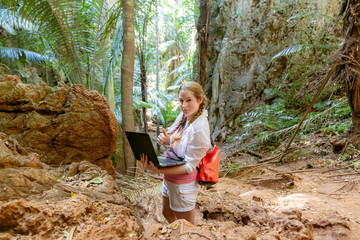 Young woman in white works with a laptop in the mountains and tropical jungle. Work in travel. He looks into the camera. Freelancer at work. Showing thumbs up