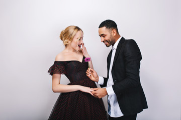 Handsome man in tuxedo giving flower to beautiful young blonde woman in luxury evening dress on white background. Astonished, joyful, happy moments, Valentines day, gift, love, together