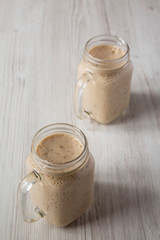 Glass jar mugs filled with banana, kiwi, apple smoothie on a white wooden background, low angle view.