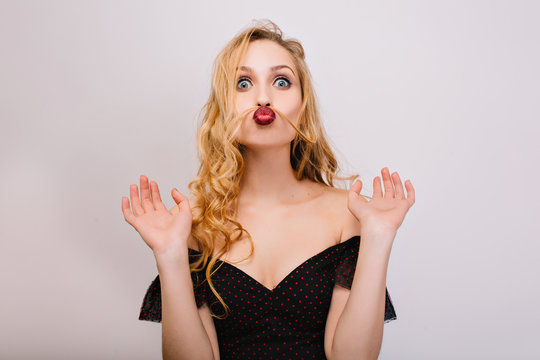 Closeup portrait of funny blonde girl being crazy, having fun, making faces, imitating moustache with hair. She has beautiful curly hair, red lips. Wearing black dress. Isolated.
