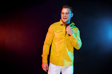 Positive young male looking away and adjusting trendy yellow shirt while standing under colorful light during party