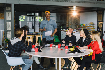 African american man in casual wear standing near the glass office wall with financial points, explaining planning and business strategies to colleagues, creative solutions on brainstorming session