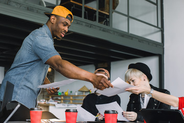Meeting Talking Discussion Brainstorming Communication Concept. Young handsome African man in jeans shirt giving papers for his coworkers in modern office during meeting