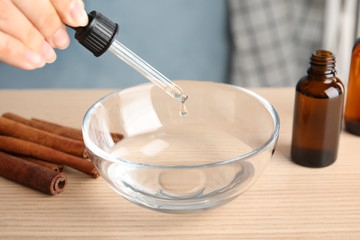Woman dripping cinnamon essential oil from pipette into bowl of water on table, closeup