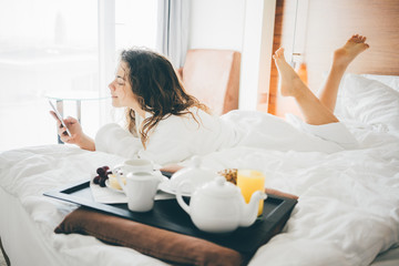 Woman having breakfast in the hotel room and looking to the phone. . Tray with breakfast on a bed.