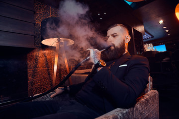 Pensive tattooed man is relaxing on the armchair and is smoking hookah, making hazy vapour.