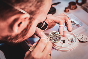 Mature clockmaster is fixing old watch for a customer at his busy repairing workshop.