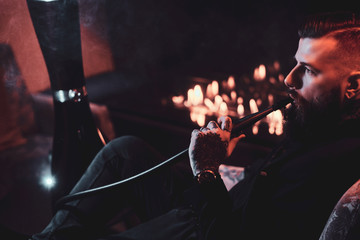 Groomed bearded man is relaxing on lounge near fireplace while smoking hookah. He has tattoo on his...