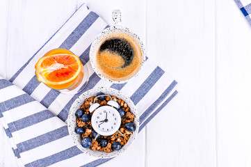 Muesli with blueberries in a cup, orange juice and aromatic morning coffee. Breakfast on a white wooden background and an alarm clock. Flat lay. Copy space.