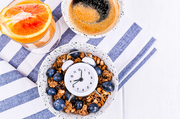 Muesli with blueberries in a cup, orange juice and aromatic morning coffee. Breakfast on a white wooden background and an alarm clock. Flat lay. Copy space.