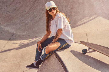 Beautiful young woman in sunglacces and cap is sitting at skatepark on the ramp with her longboard. 